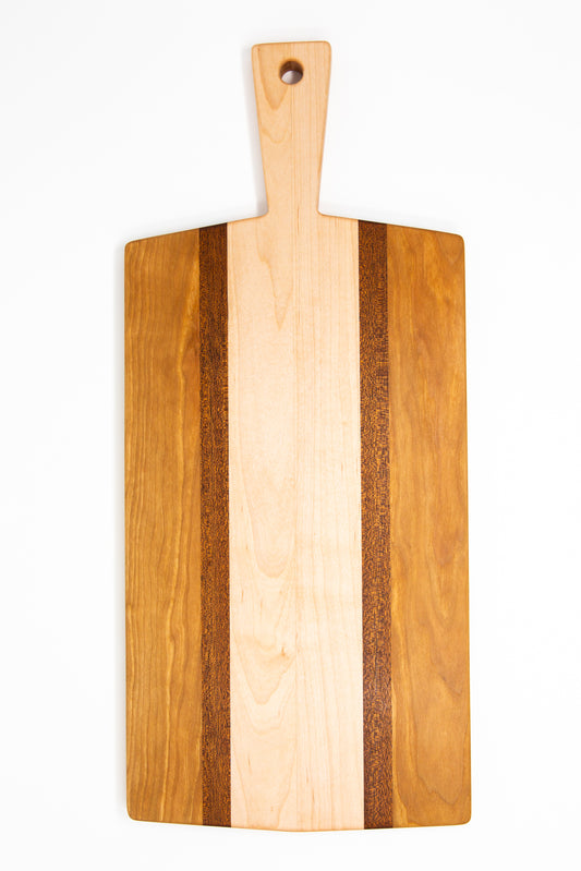 Large size wood charcuterie board, made of golden-brown Cherry, reddish brown Sapele and a center piece of light blonde Maple with a slightly tapered handle and a long rectangular body.
